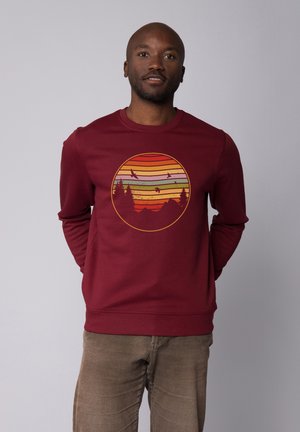 Sweatshirt bordeaux avec un motif circulaire comprenant des rayures horizontales orange, verte et jaune, représentant des arbres et des montagnes.