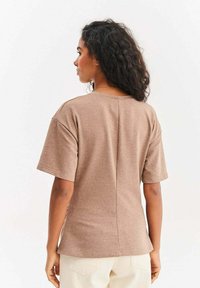 Kobieta o kręconych, ciemnych włosach, ubrana w brązową koszulkę z krótkim rękawem i kremowe spodnie, zwrócona tyłem na białym tle.