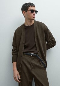 Brown button-up cardigan over a dark t-shirt, paired with brown trousers. The model wears black sunglasses and a braided belt.