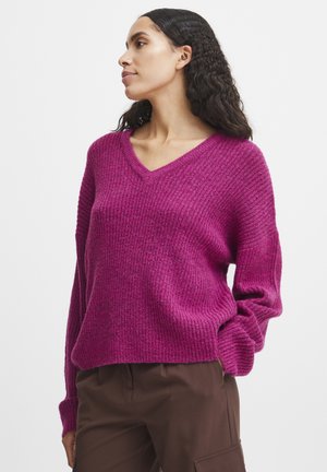 V-Ausschnitt Pullover in leuchtendem Fuchsia, mit gerippter Textur und langen Ärmeln, kombiniert mit braunen Hosen.
