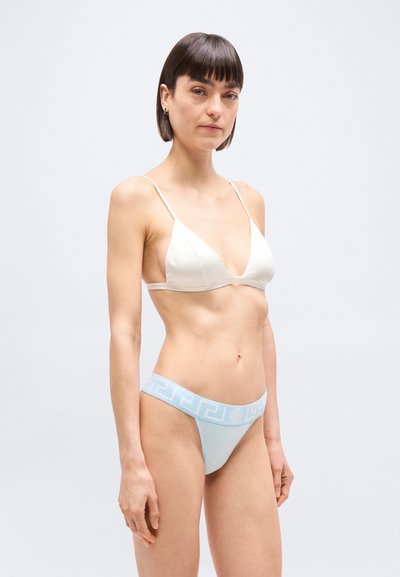 Γυναίκα που φοράει λευκό σατέν τρίγωνο σουτιέν και ανοιχτό μπλε εσώρουχα με ζώνη στη μέση με μοτίβο Ελληνικού κλειδιού, στέκεται μπροστά σε απλό λευκό φόντο.