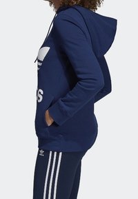 Navy blue collegehuppari, jossa on suuri valkoinen logo, pitkät hihat ja etutaskut; yhdistettynä tummansinihin leggingseihin, joissa on kolme valkoista raitaa.