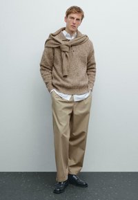 Beige gestrickter Pullover mit Rundhalsausschnitt, darunter ein hellblaues Hemd mit Kragen, kombiniert mit lockeren beigen Hosen und schwarzen Slippern.