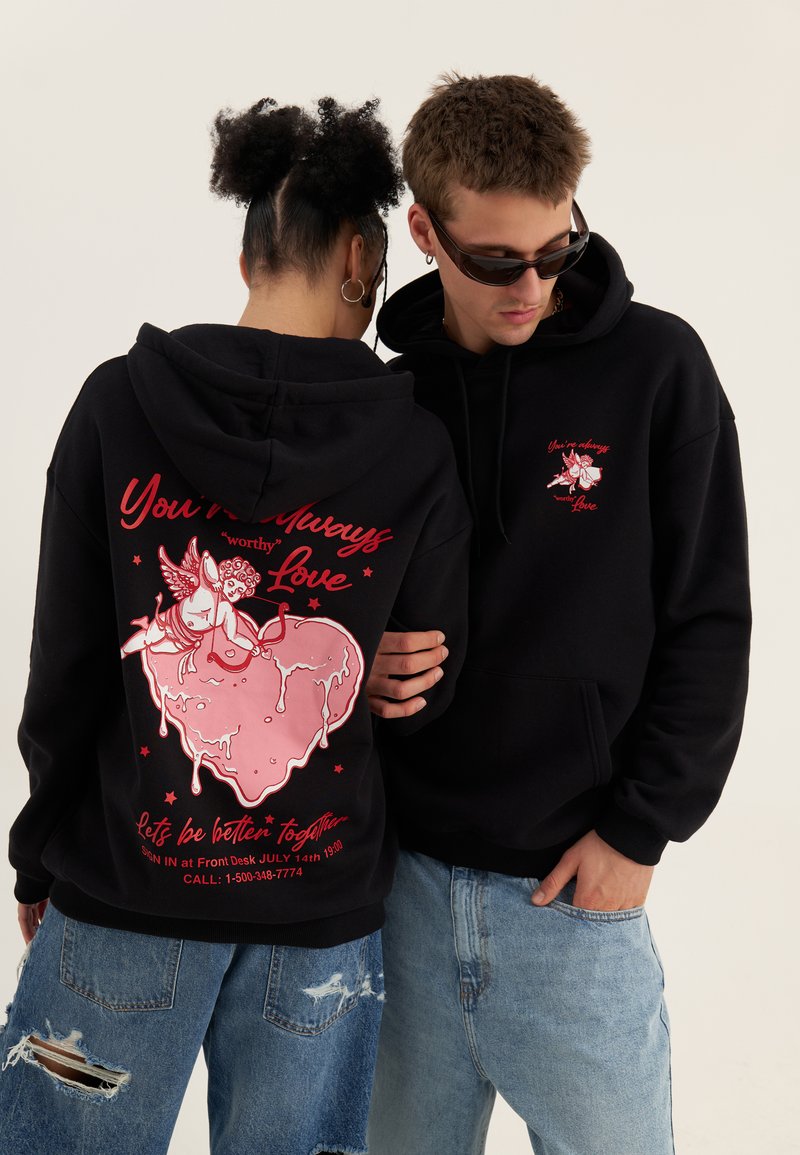 Two people wearing black hoodies with pink cupid and heart graphics and red text, embracing; both wear light blue ripped jeans.
