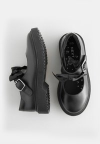 Souliers Mary Jane en cuir noir avec un bout arrondi, semelle épaisse texturée, sangle à boucle réglable et accent de nœud sur le dessus.