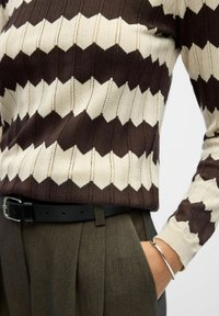 Braun- und cremefarbener Zickzackmuster-Strickpullover kombiniert mit grauen, gefalteten Hosen und einem schwarzen Gürtel. Ein silbernes Armband schmückt das Handgelenk.