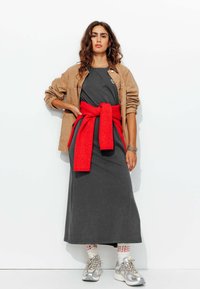 Robe longue grise avec une ceinture en tricot rouge nouée, associée à une veste beiger et des baskets argentées. Tissu texturé, design décontracté et look superposé.
