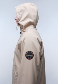 Beige vattentät jacka med huva, prydd med en cirkulär märkespatch på vänster ärm och kontrasterande sömmar. Slät, matt yta.