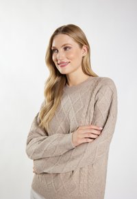 Pulover bej din tricot cu modele în diamant, mâneci raglan și manșete cu elastic. Textură moale și croială lejeră, purtat cu brațele încrucișate.