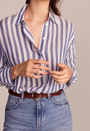 Frau trägt ein blau-weiß gestreiftes Hemd mit Knöpfen, das in hoch taillierte blaue Jeans gesteckt ist, dazu einen braunen geflochtenen Gürtel.