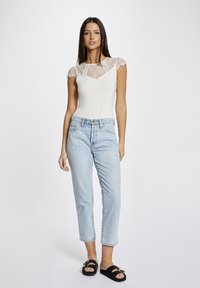 White top with lace cap sleeves, V-neck, paired with light blue high-waisted jeans and black slide sandals with decorative hardware.