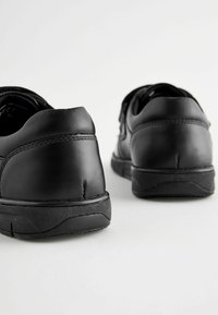 Chaussures en cuir noir avec une texture lisse et des talons arrondis. Les semelles sont en caoutchouc et les coutures sont visibles le long des bords.