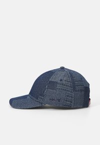Sötétkék farmerből készült baseballsapka, foltvarrásos mintázattal, eltérő textúrákkal és ívelt napellenzővel. A hátoldalon logó részlet található.