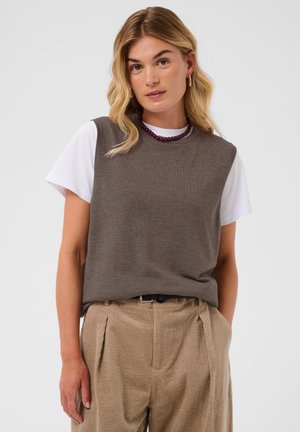 Jeune femme aux cheveux blonds portant un pull sans manches marron par-dessus un t-shirt blanc et un pantalon en velours côtelé beige plissé, debout, les mains dans les poches.