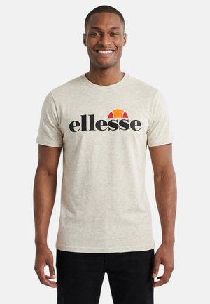 Homme souriant portant un t-shirt beige clair avec un texte noir « ellesse » et un logo demi-cercle orange-rouge, assorti à un pantalon noir.
