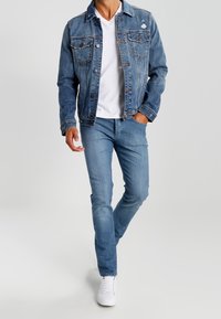 Džínová bunda s vybledlým modrým odstínem, dvěma předními kapsami a jemným opotřebením. Nošená přes bílé tričko a kombinovaná s světle modrými džíny.