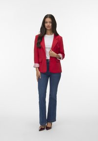 Blazer rouge avec un seul bouton, haut blanc et jeans évasés bleus. Détails de poignets rayés et escarpins bordeaux à bout pointu.