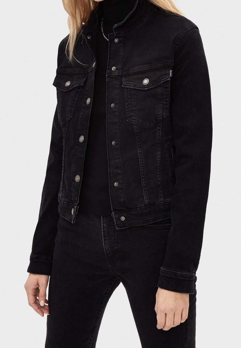 Mustan denimin takki, jossa on napitettu etuosa, kaksi rintataskua ja istuva muotoilu. Kankaan pinnassa on hienovarainen rakenne ja himmeä viimeistely.