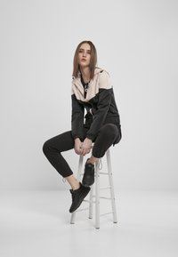 Svart og beige vindjakke med glidelås foran, elastiske mansjetter og justerbar hette, kombinert med svarte joggebukser og svarte sneakers på en krakk.