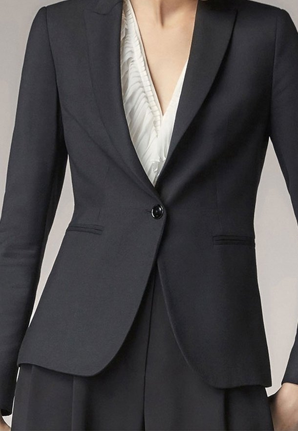 Femme portant un blazer noir cintré sur un chemisier blanc à volants, boutonné à la taille, avec un pantalon noir assorti.