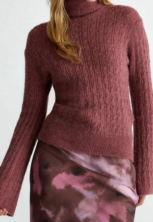 Kvinde iført en tekstureret mauve rullekravesweater og et skinnende, brunt og pink batikfarvet skørt, vist fra bryst til hofter.