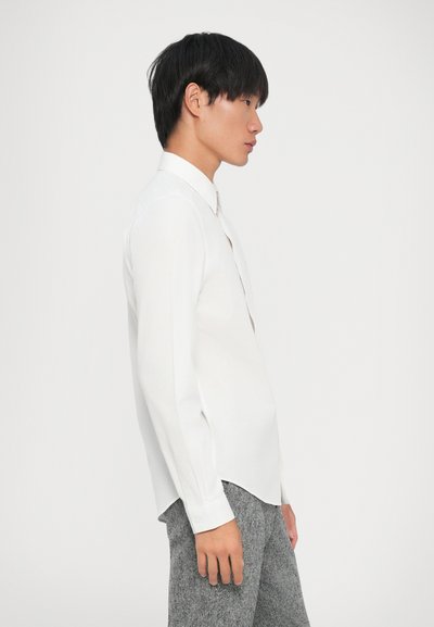 Camicia bianca a maniche lunghe con colletto classico, tessuto morbido, spacchi laterali e polsini con bottoni, abbinata a pantaloni grigi a trama.