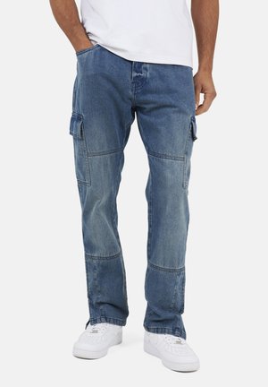Mężczyzna w niebieskich dżinsowych spodniach typu cargo z klapkowymi kieszeniami i białych tenisówkach, stojący z jedną ręką w kieszeni.