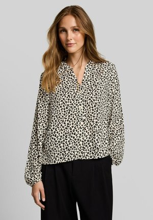 Frau mit langen Haaren, die eine cremefarbene Bluse mit schwarzen Leopardenflecken und schwarze Hose trägt, vor einem einfarbigen grauen Hintergrund stehend.