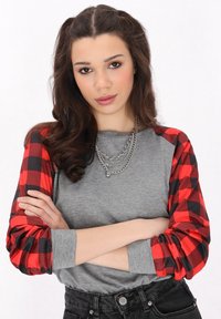 Grå långärmad topp med röda och svarta rutiga ärmar, kombinerad med svarta högmidjade jeans och lager av silverbroscher.