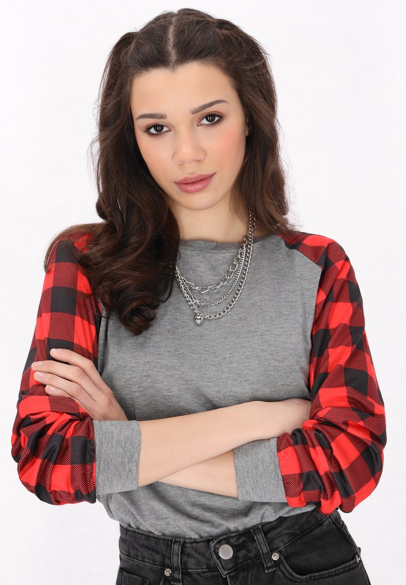 Grå långärmad topp med röda och svarta rutiga ärmar, kombinerad med svarta högmidjade jeans och lager av silverbroscher.