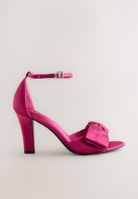 Vaaleanpunainen satiininen korkokengänkengännauha, jossa on leveä remmi, säädettävä nilkkahihna hopeisella soljella ja suuri rusetti edessä.