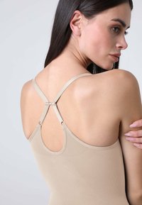 Top beige senza maniche con un design a tracolla incrociato, dotato di hardware regolabile e una trama liscia e elastica. La vista posteriore mostra una corrispondenza con il tono della pelle.