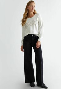 Maglione bianco con motivo geometrico di perline, abbinato a jeans neri a gamba larga con vita alta. Il modello indossa scarpe con punta appuntita e decorazioni.