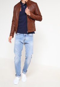 Brun läderjacka med ståkrage, parat med en marinblå polo, ljusblå jeans och vita sneakers.