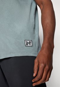 Grågrøn bomulds tanktop med et sort og hvidt Nike-logo patch ved kanten. Glat tekstur, afslappet pasform, parret med sorte shorts.