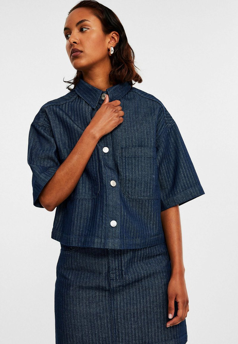Korte denim shirt in donkerblauw met verticale strepen, korte mouwen en een knoopsluiting. Bevat twee voorkruiden en zilverkleurige knopen.