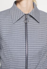 Jas met rits met een geometrisch patroon in marineblauw en wit, een gestructureerde kraag en een cirkelvormig hardware-detail aan de rits.