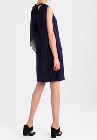 Robe sans manches bleu marine avec un superposé fluide en forme de cape, maintenu au cou par des liens. Associée à des sandales à talons carrés noires.
