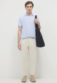 Jeune homme portant un polo bleu clair à manches courtes, un pantalon crème, des chaussures beiges à enfiler, et portant un sac à épaule noir.