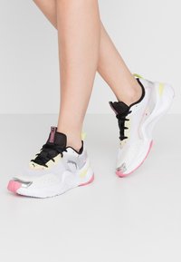 Weiße Sportschuhe mit schwarzen Akzenten, einem Obermaterial aus Mesh, silbernem Detail und einer pinken sowie gelben Sohle, ausgestattet mit Schnürsenkeln und einem gepolsterten Kragen.