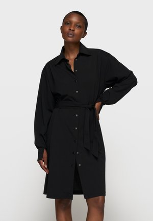 Donna che indossa un vestito camicia nero a maniche lunghe con cintura annodata in vita, in piedi davanti a uno sfondo chiaro e semplice.