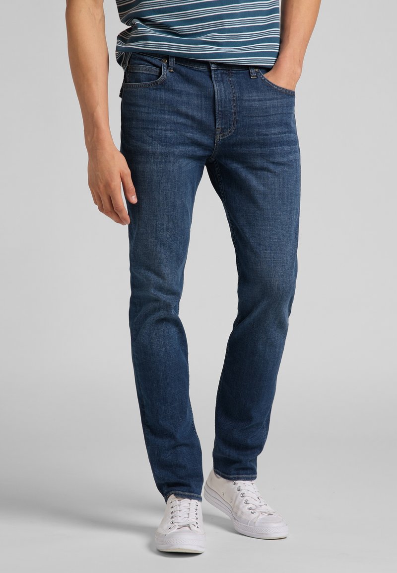 Man som bär slim fit mörkblå jeans, vita sneakers och en randig skjorta med en hand i fickan mot en grå bakgrund.