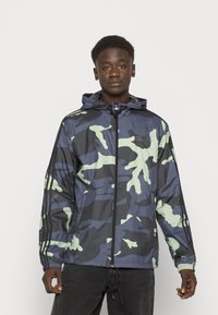 Giacca a vento mimetica in blu navy e verde chiaro, con cappuccio, chiusura con zip e polsini elastici. Realizzata in materiale leggero e lucido.