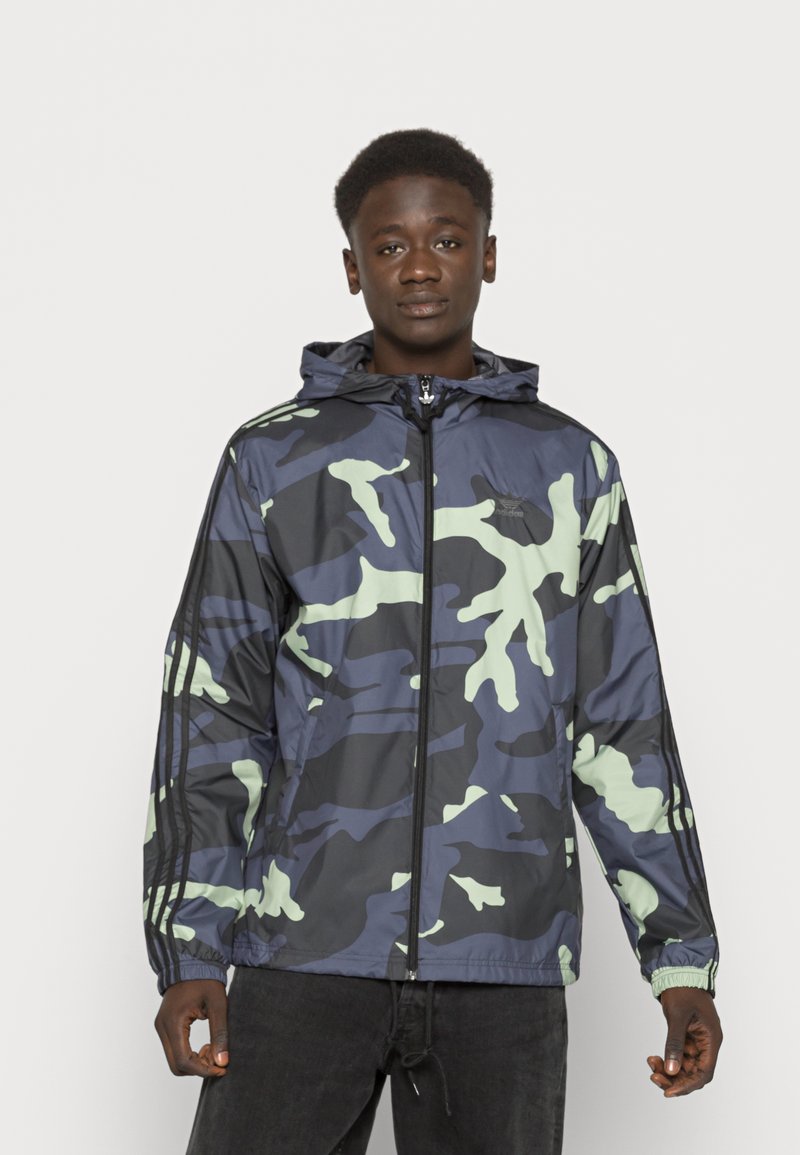 Giacca a vento mimetica in blu navy e verde chiaro, con cappuccio, chiusura con zip e polsini elastici. Realizzata in materiale leggero e lucido.