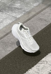 Chaussure de sport blanche avec une tige en mesh pour la respirabilité, semelle rembourrée, accents texturés sur les côtés et un design de laçage sécurisé.