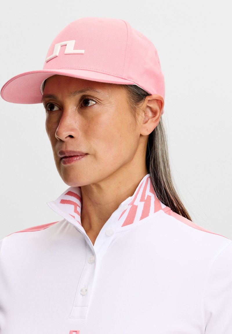 Donna che indossa un berretto rosa con logo bianco e una camicia bianca con colletto, decorata con motivi rosa sul colletto e sulle spalle.