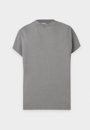 Femme aux cheveux courts portant un haut gris sans manches ample et un pantalon large bleu marine, debout une main dans la poche, sur un fond uni.