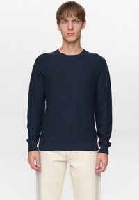 Jonge man met kort bruin haar, gekleed in een marineblauwe gebreide trui en roomwitte broek, staand tegen een eenvoudige lichte achtergrond.