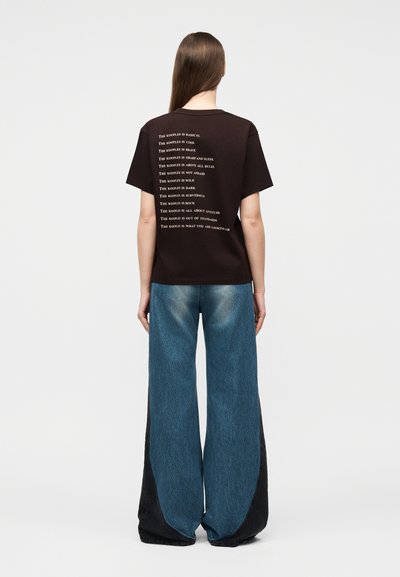 Άτομο με μακριά καστανά μαλλιά φοράει μαύρο μπλουζάκι με λευκό κείμενο και μπλε τζιν παντελόνι με φαρδιά πόδια και μαύρες πλαϊνές ραφές, στέκεται με την πλάτη προς τα εμπρός.