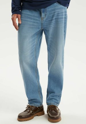 Mężczyzna w jasnoniebieskich dżinsach i brązowych skórzanych butach sznurowanych, stoi z jedną ręką swobodnie opuszczoną wzdłuż ciała.